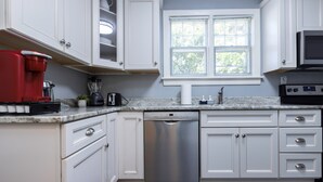 Una cocina con armarios blancos, lavavajillas, cafetera roja y ventana con persianas.