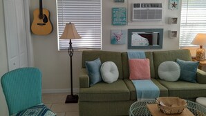 A cozy living room with a green sofa, a turquoise armchair, a guitar, and various wall decorations including a 'Welcome' sign and a 'This is my happy place' sign.