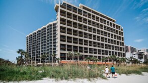 Un hotel de varios pisos con fachada enrejada, situado cerca de una playa con palmeras y arena.