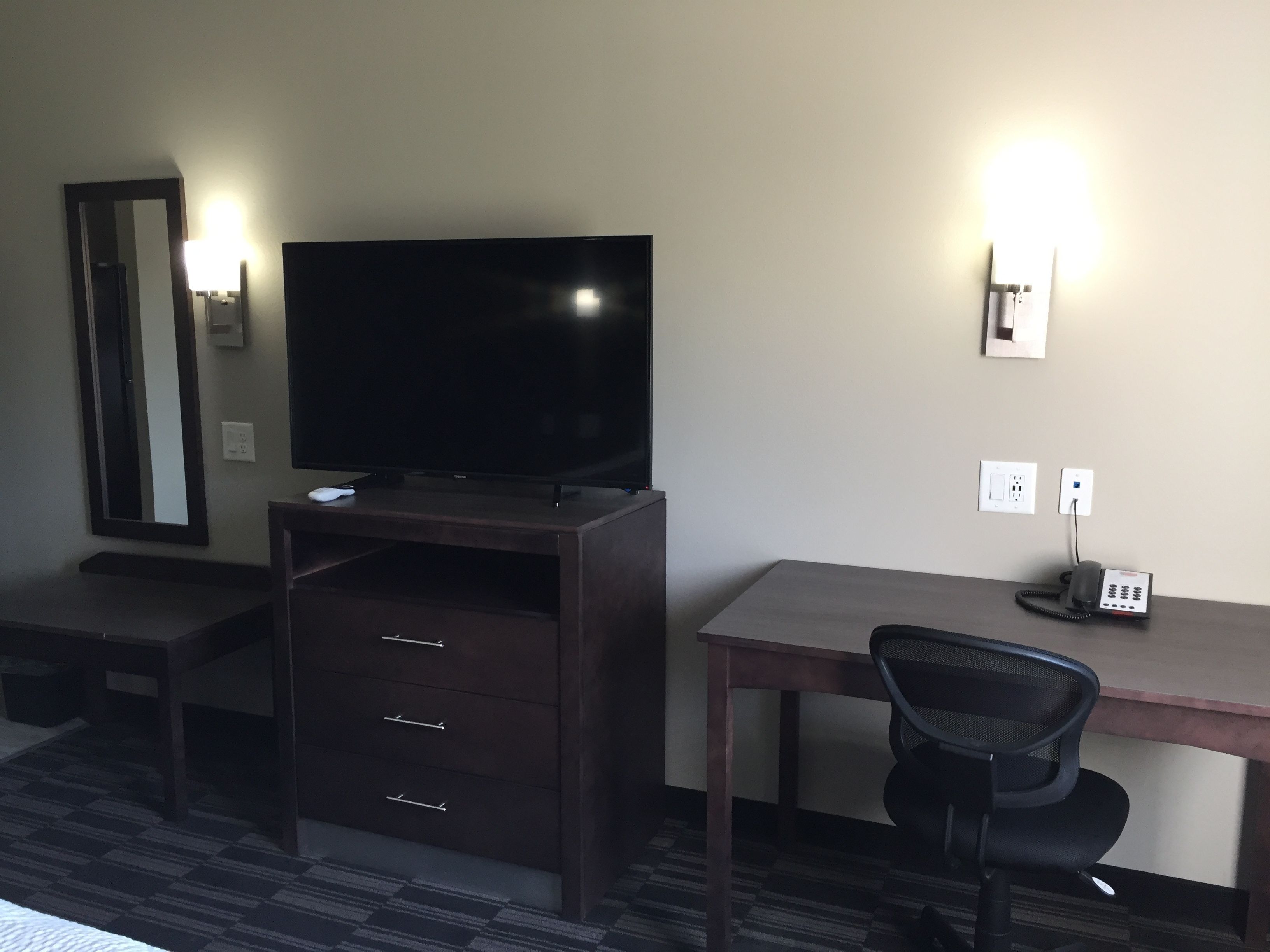 A hotel room with a flat-screen TV on a wooden cabinet, a desk with a chair, and a phone on the desk.