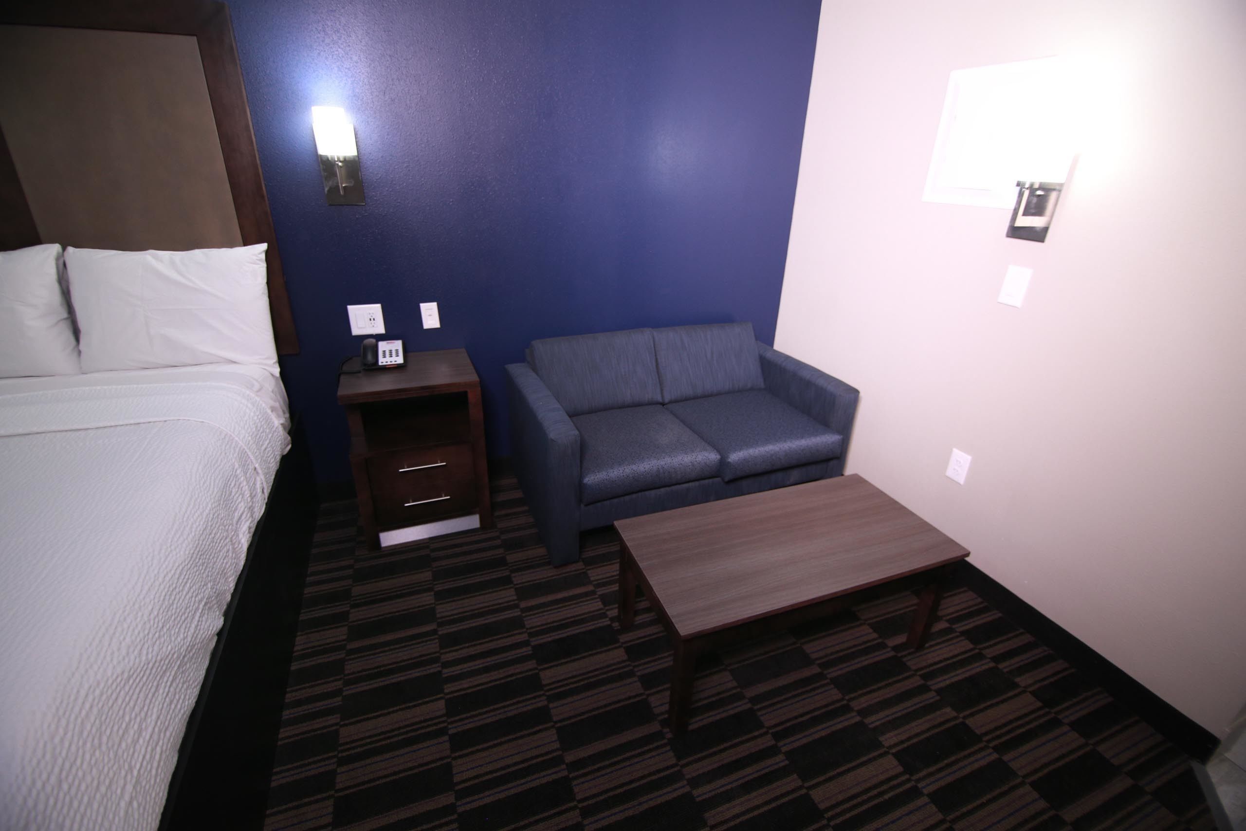 A hotel room with a bed, a gray sofa, a small wooden table, and a carpeted floor.