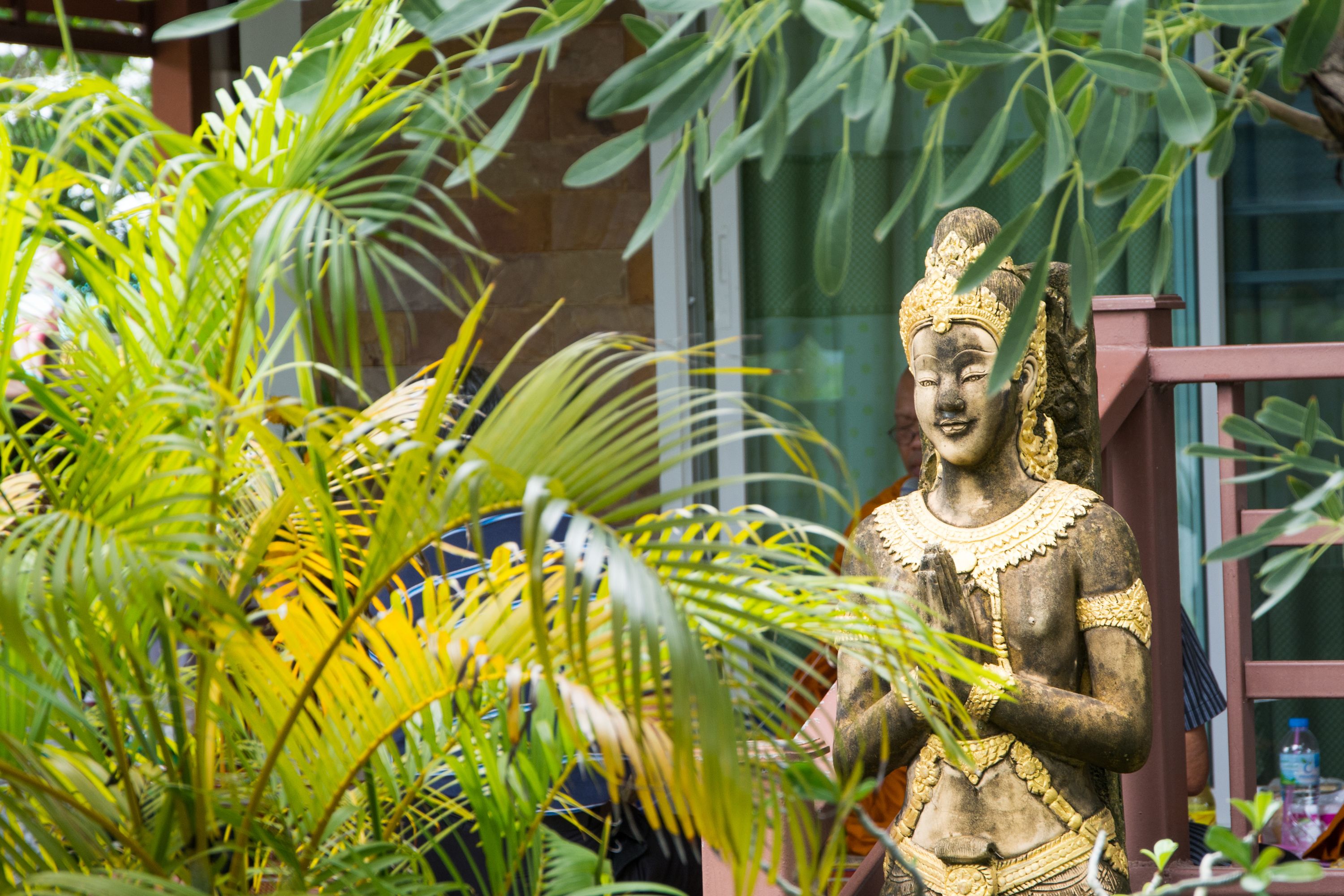 A statue of a figure with hands in a prayer position, surrounded by lush green foliage.