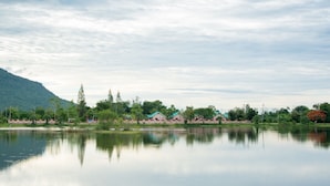Un lago tranquilo con el reflejo de edificios y árboles, rodeado de vegetación y una montaña al fondo.