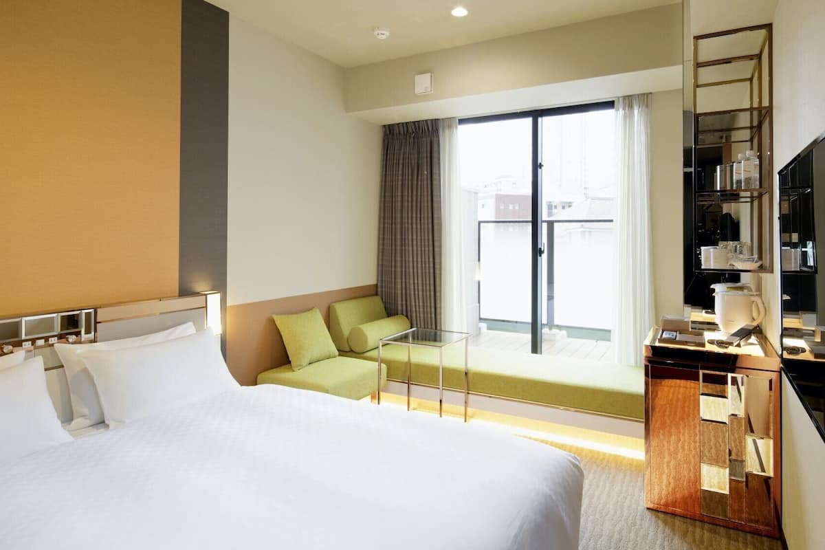 A hotel room with a large bed, a seating area with a glass table, a balcony with curtains, and a minibar.