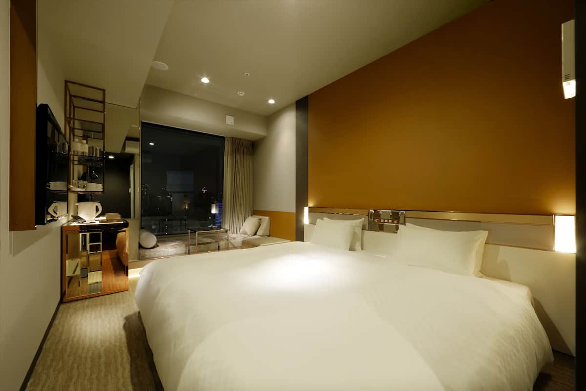 A hotel room with a large bed, a desk with a mirror, a chair, and a view of the city at night.