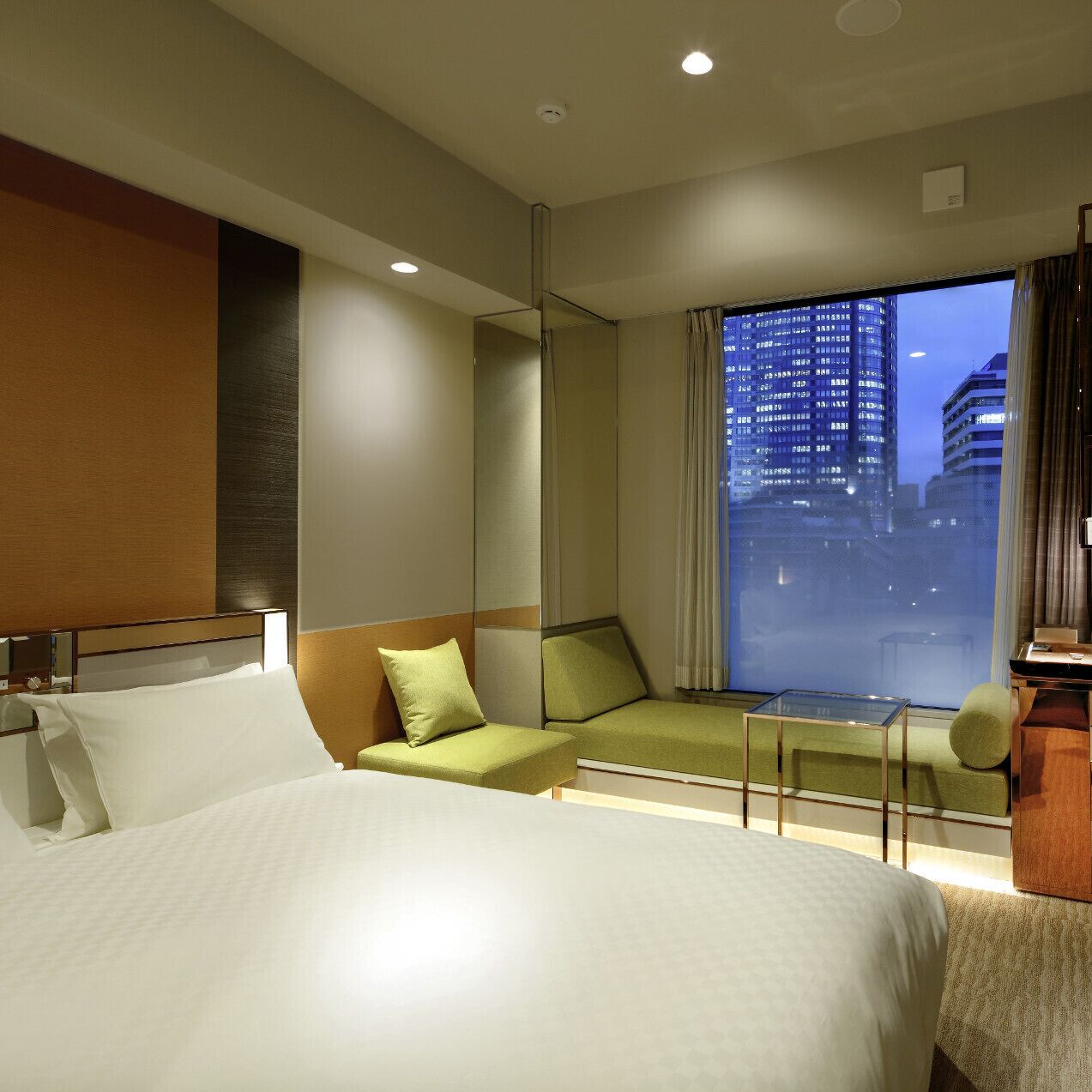 A hotel room with a bed, a green sofa, a small table, and a view of a cityscape at night.