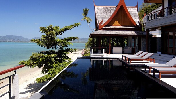 An infinity pool with lounge chairs, a beach view, and a traditional building with a pointed roof.