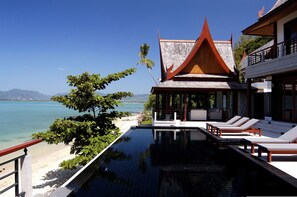 Une piscine à débordement avec des chaises longues, une vue sur la plage et un bâtiment traditionnel doté d’un toit en pente.
