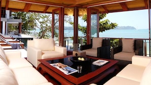 A spacious indoor area with a wooden ceiling, large windows offering a view of the sea, and a seating area with white sofas and a coffee table.