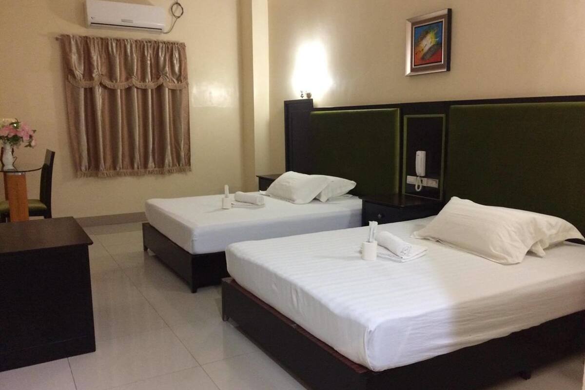 A hotel room with two single beds, a desk, a chair, an air conditioner, a telephone, and a painting on the wall.