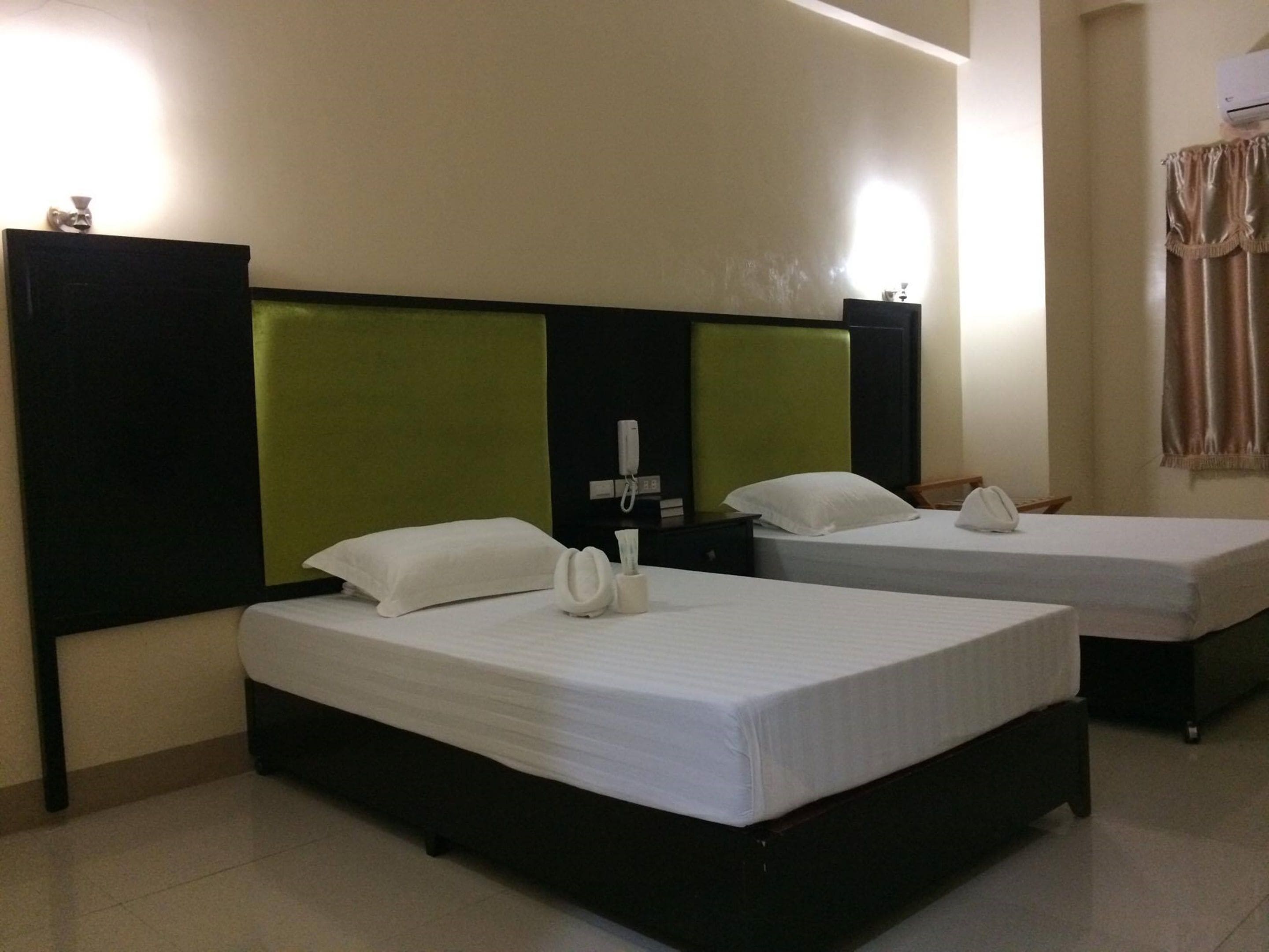 Quarto de hotel com duas camas de solteiro, cada uma com colcha e travesseiros brancos, cabeceira verde e mesa de cabeceira com abajur e telefone.