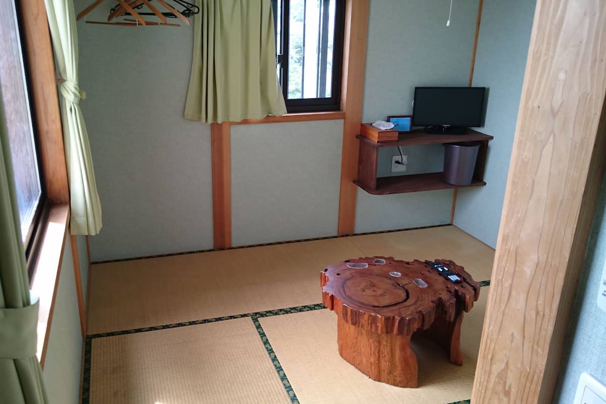 A tatami mat room with a tatami mat table, a television on a wooden shelf, and a window with curtains.