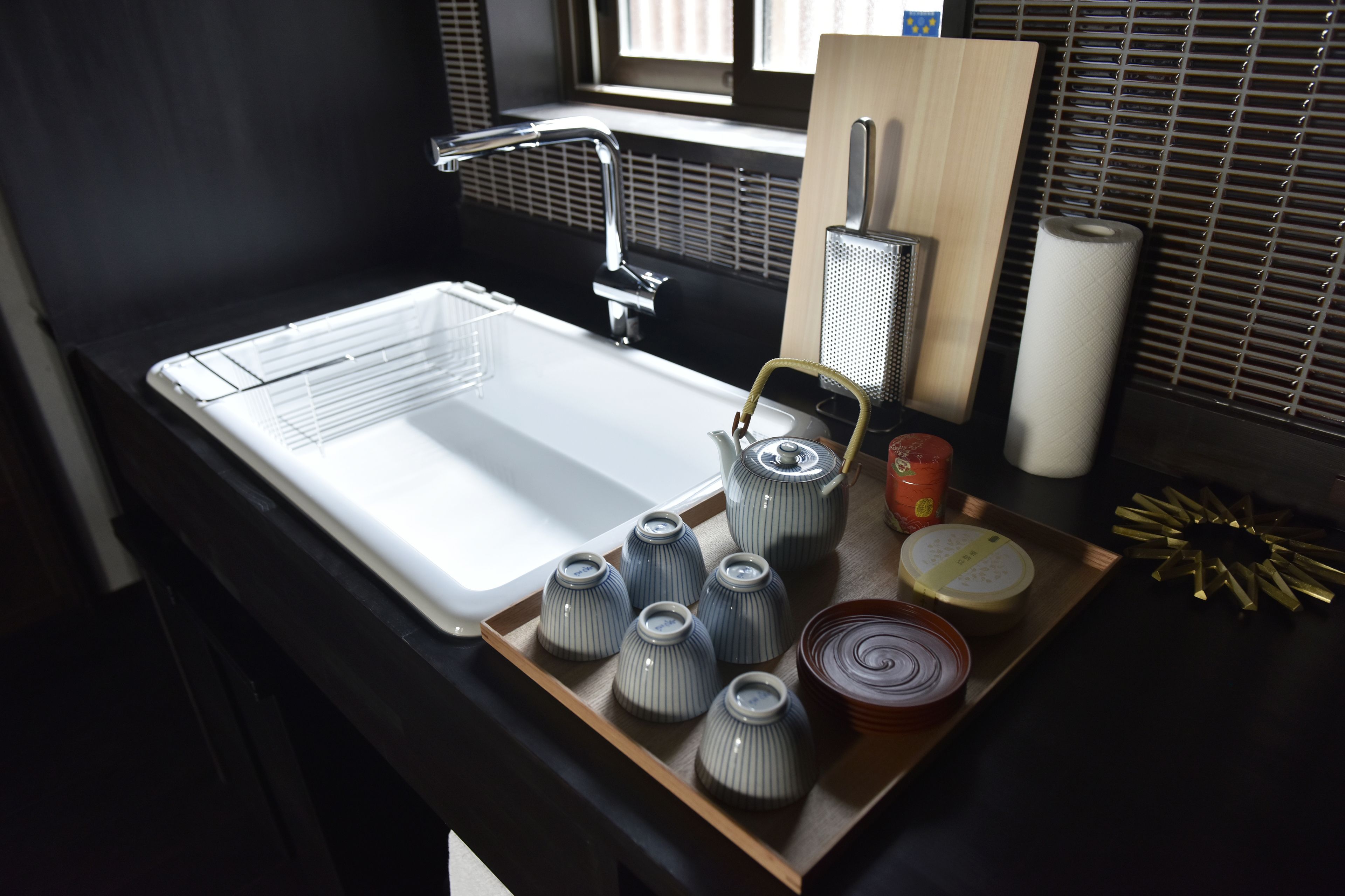 A kitchen sink with a faucet, a wooden tray with ceramic cups, a grater, a roll of paper towels, and a decorative item.