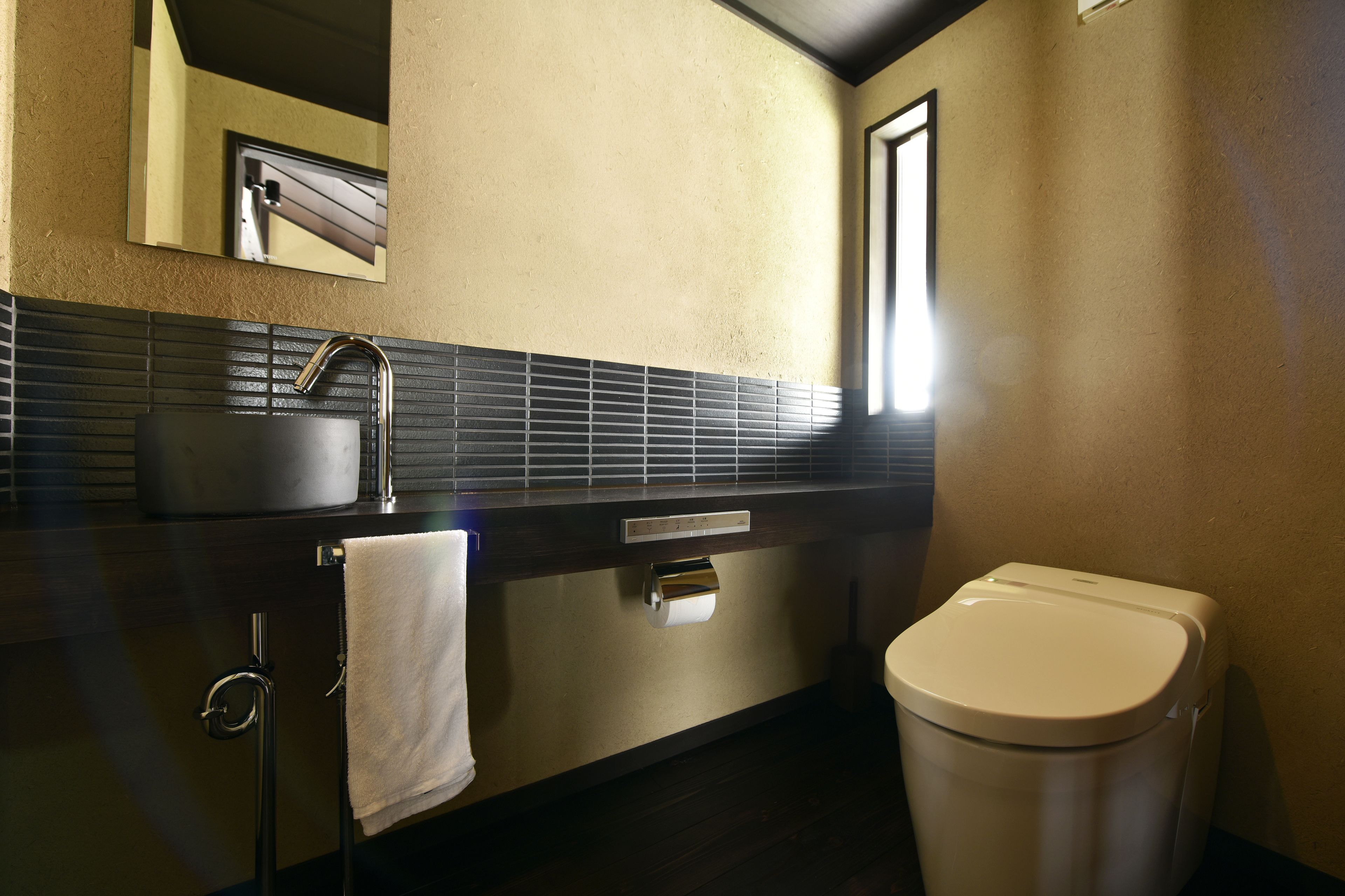 A modern bathroom with a black and beige tiled backsplash, a stainless steel sink, a toilet, and a mirror above the sink.