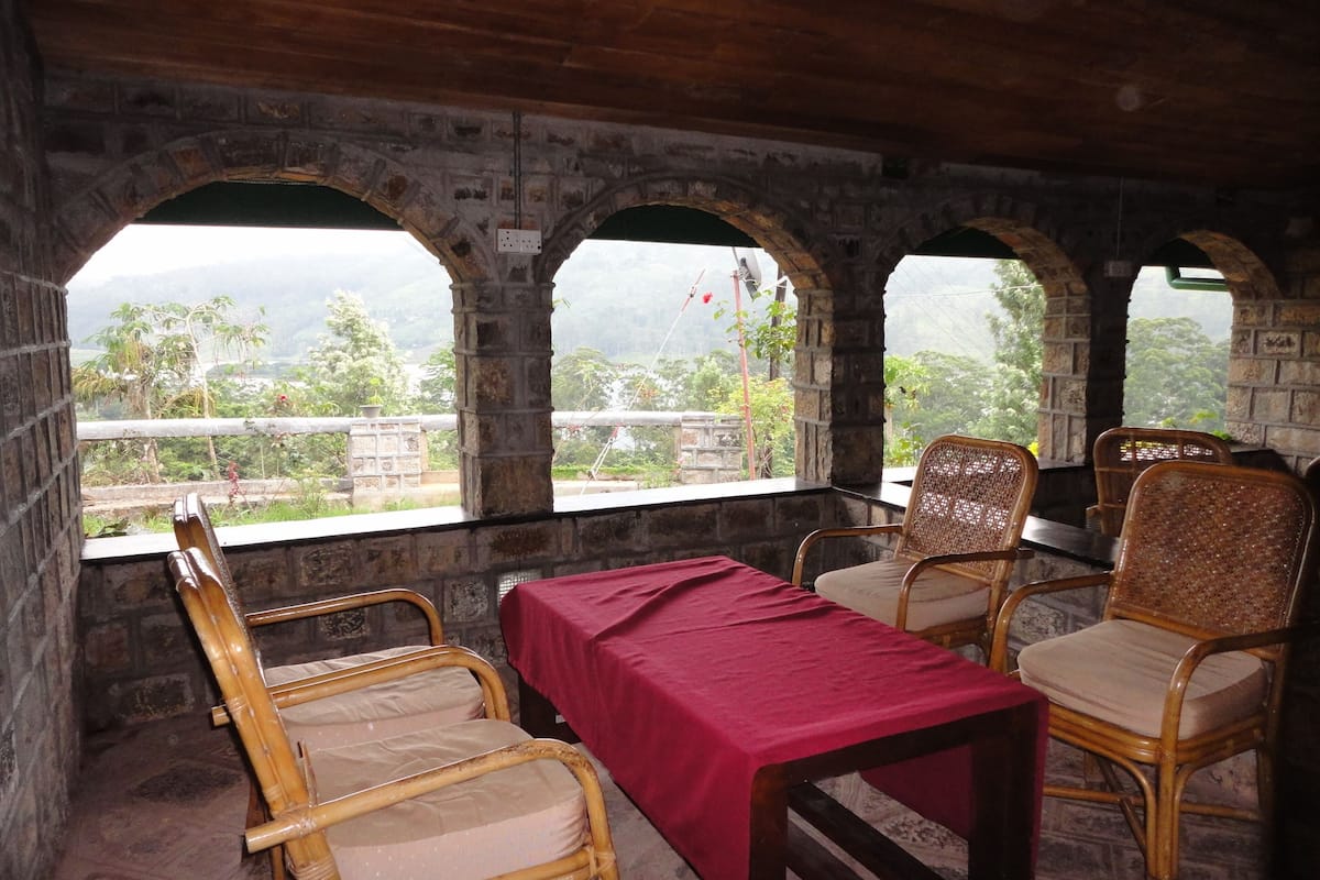 A room with a table and four chairs, a red tablecloth, and three arched windows overlooking a landscape.