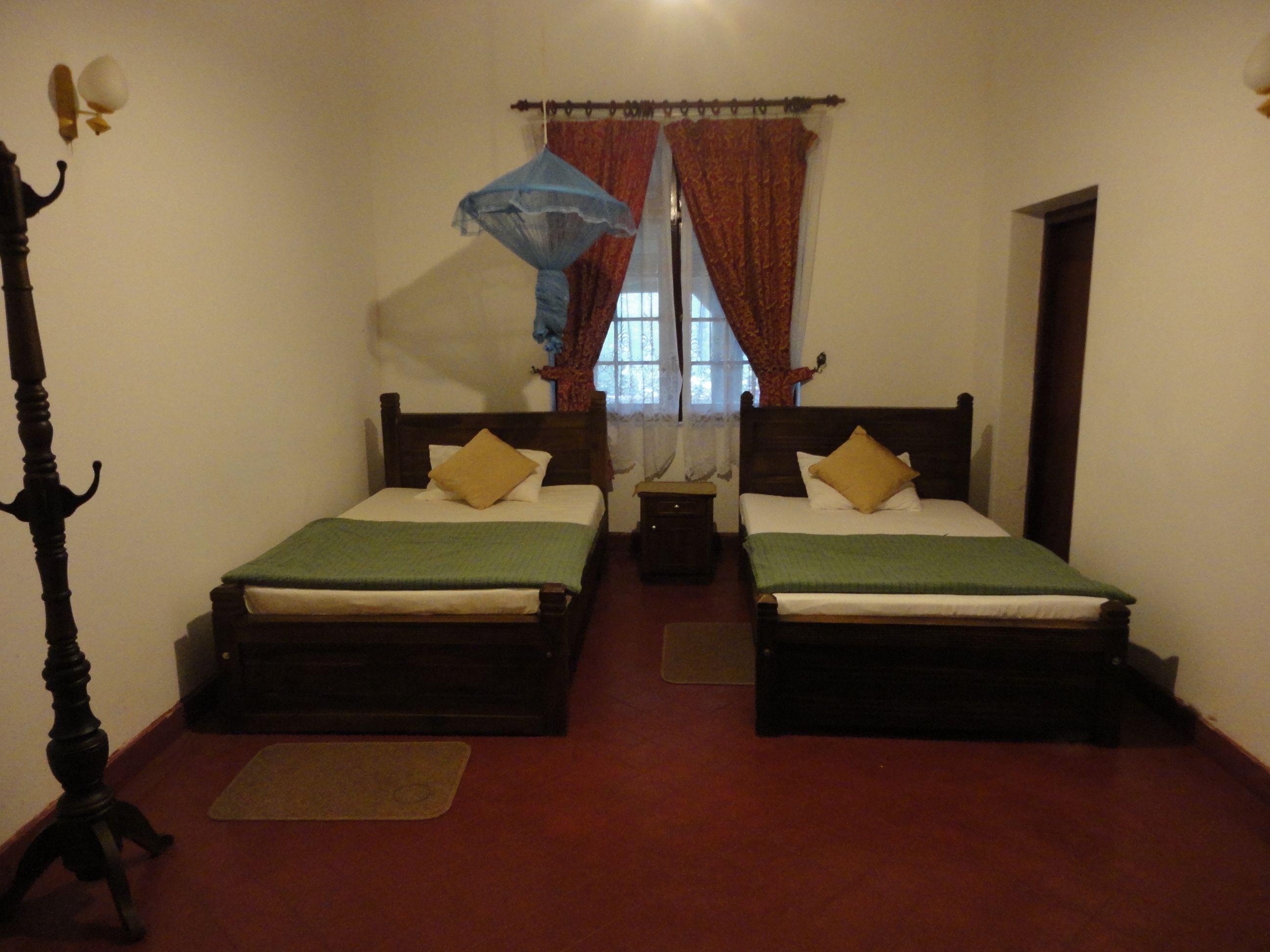 A room with two wooden beds, each with a green bedspread and a yellow pillow. There is a nightstand between the beds, and a window with red curtains and a blue mosquito net above it.