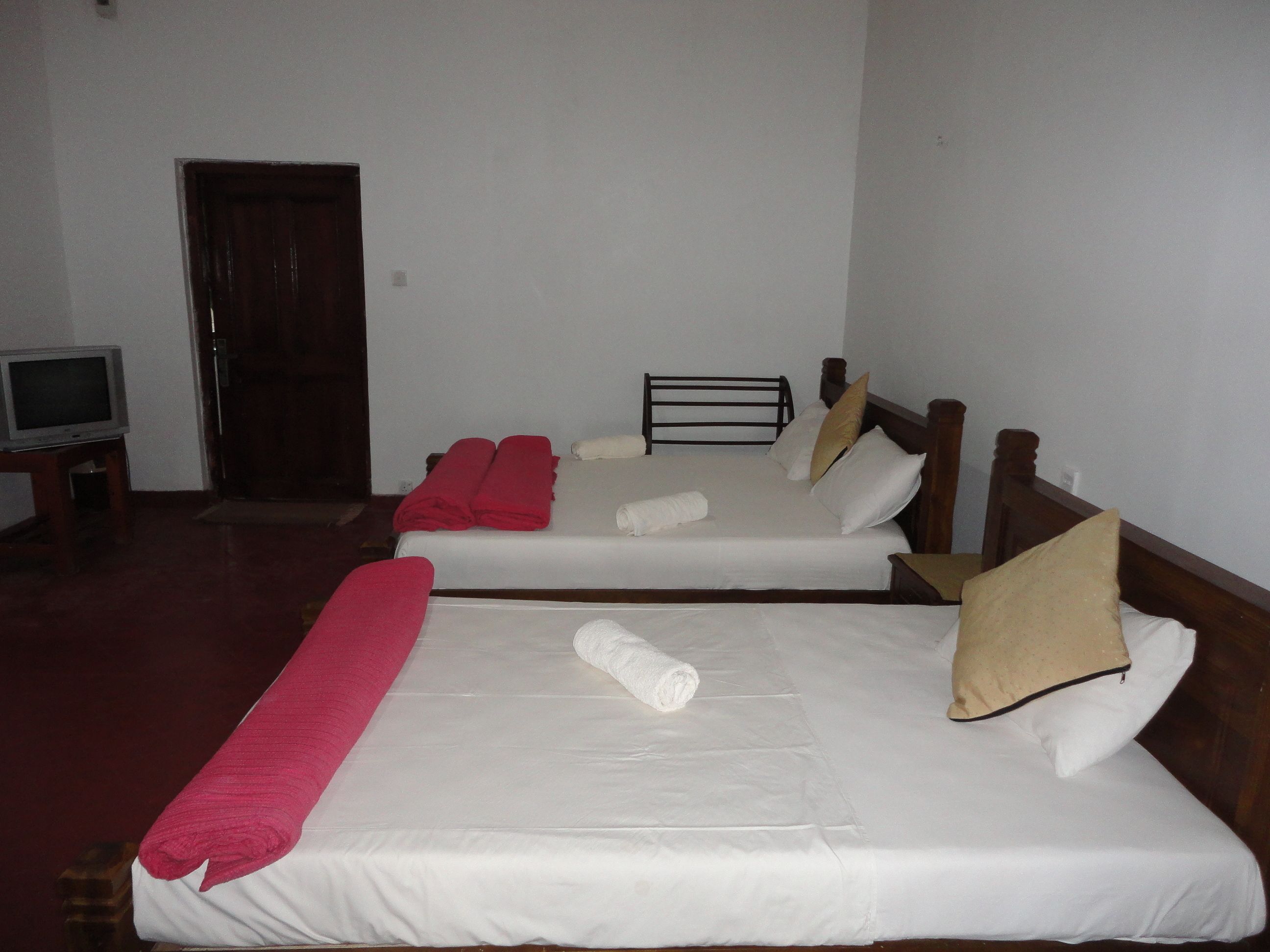 Dos camas individuales con sábanas blancas, almohadas rojas y beige, y toallas enrolladas. Una cabecera de madera está presente en una cama. Un televisor está situado sobre una mesita a la izquierda.
