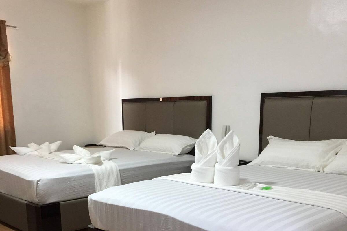A hotel room with two beds, each with white bedding and pillows. The beds have dark headboards and are placed on a wooden floor.