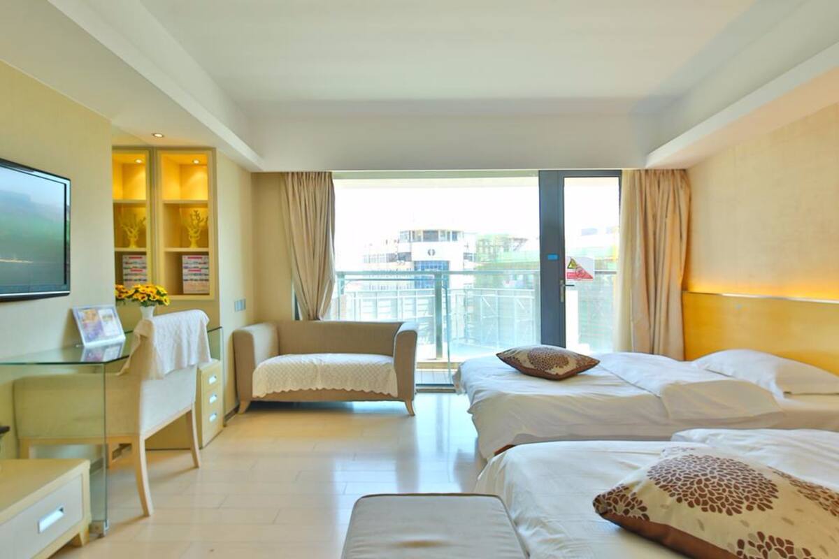 Habitación de hotel con dos camas, un escritorio con silla, televisión y balcón con vistas a edificios.