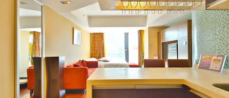 Ein modernes Hotelzimmer mit Holzboden, einer Couch, einem Bett, einer kleinen Küchenzeile und einem durch eine Glastür einsehbaren Badezimmer.