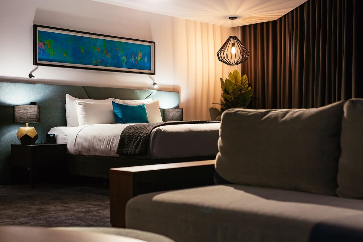 A modern hotel room with a neatly made bed, a large abstract painting, a hanging light fixture, and a potted plant.