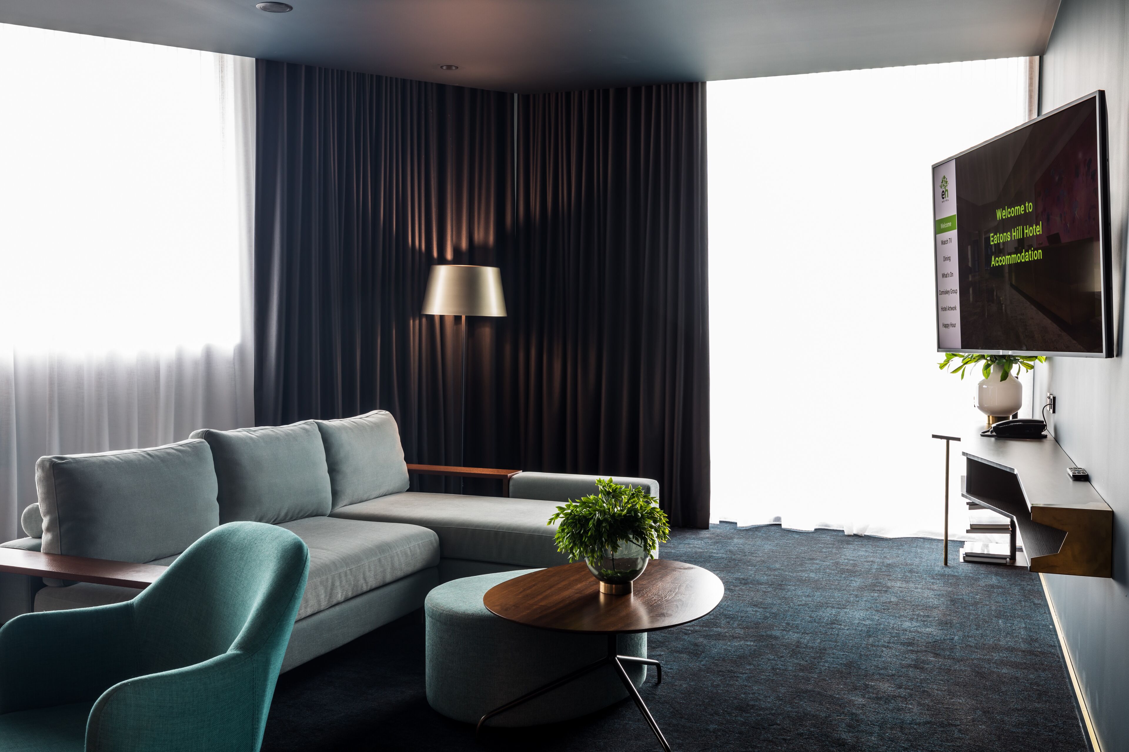 A modern hotel room with a grey sofa, a round coffee table with a plant, a TV on the wall, and a large window with curtains.