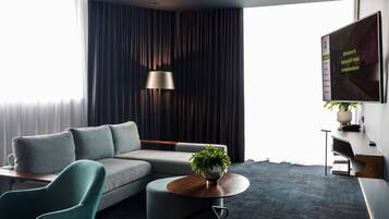 A modern hotel room with a grey sofa, a round coffee table with a plant, a TV on the wall, and a large window with curtains.