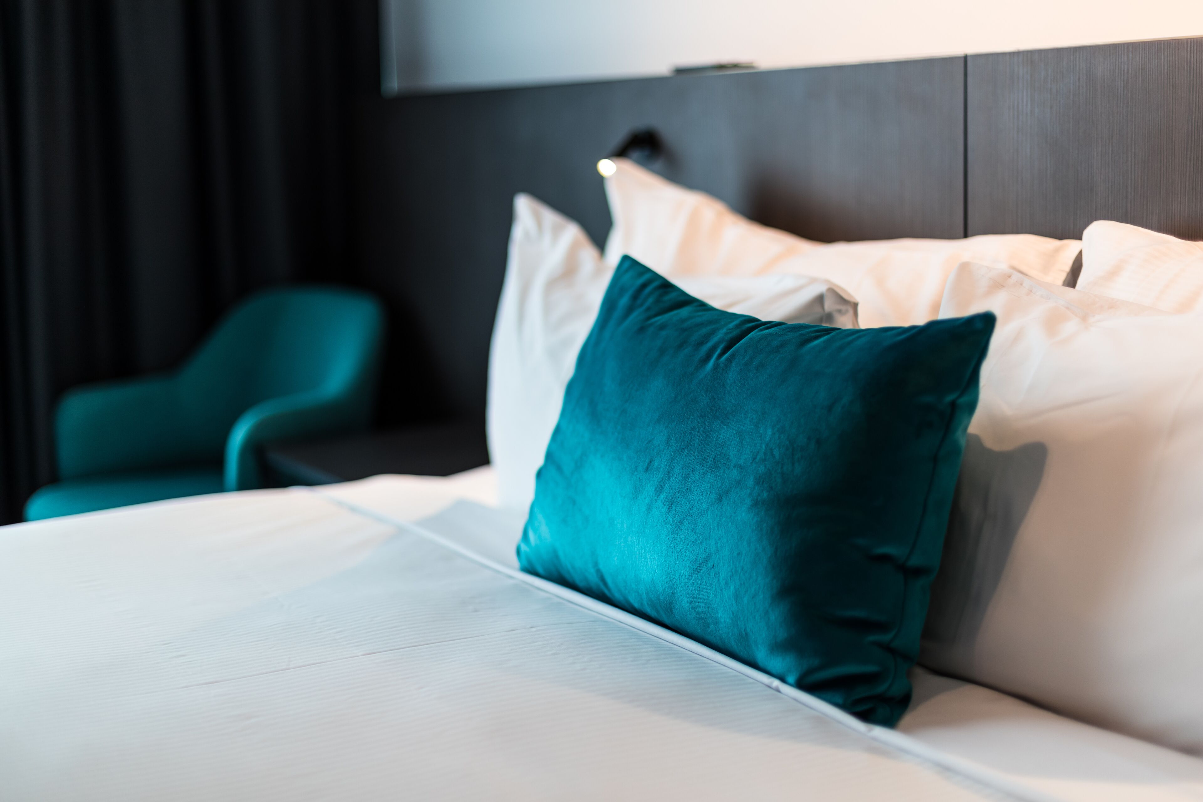 Una cama bien tendida con sábanas blancas y almohadas color aguamarina, una silla color aguamarina y un cabecero oscuro.