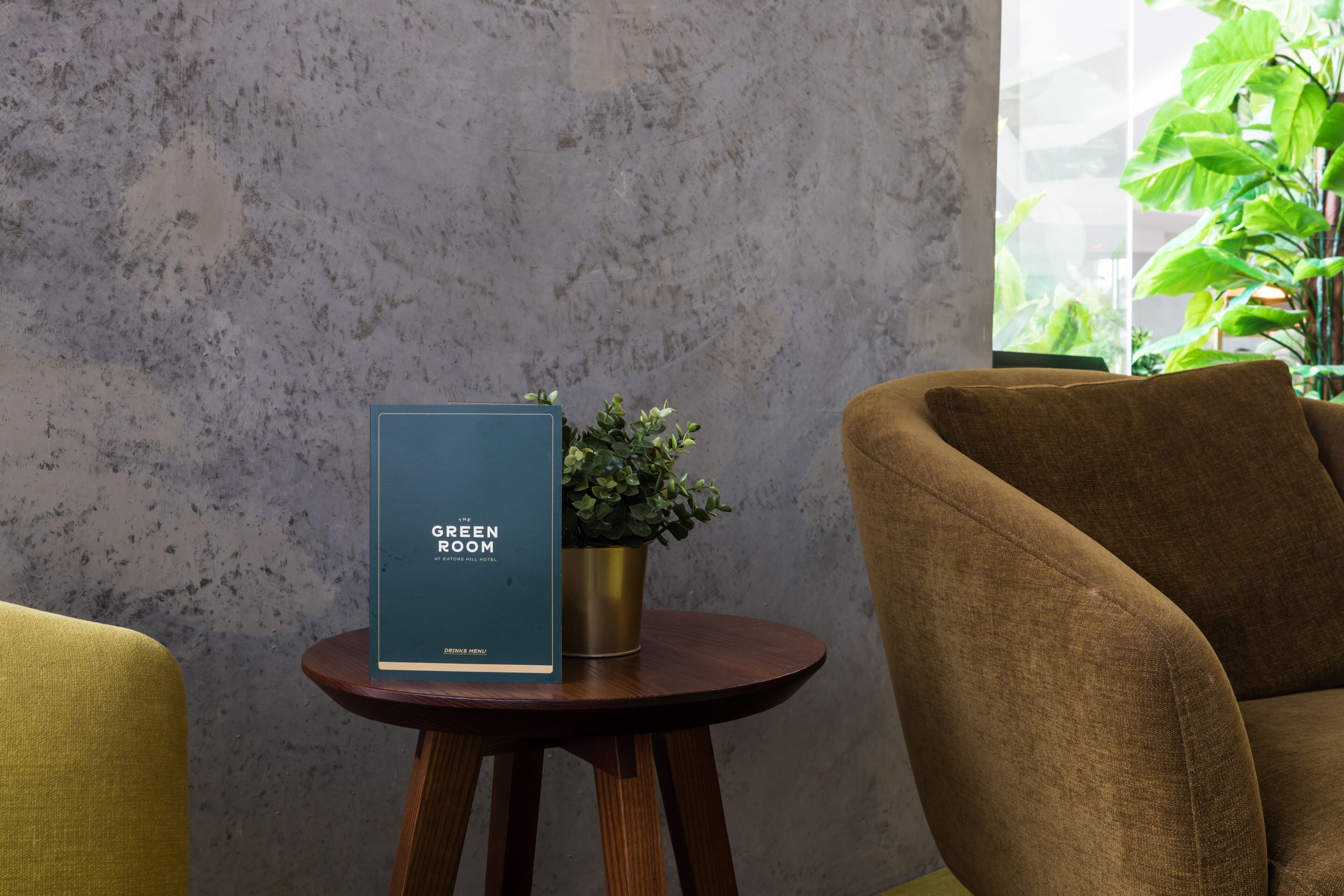 A room with a textured gray wall, a round wooden table with a blue menu stand, a potted plant, and a brown sofa.