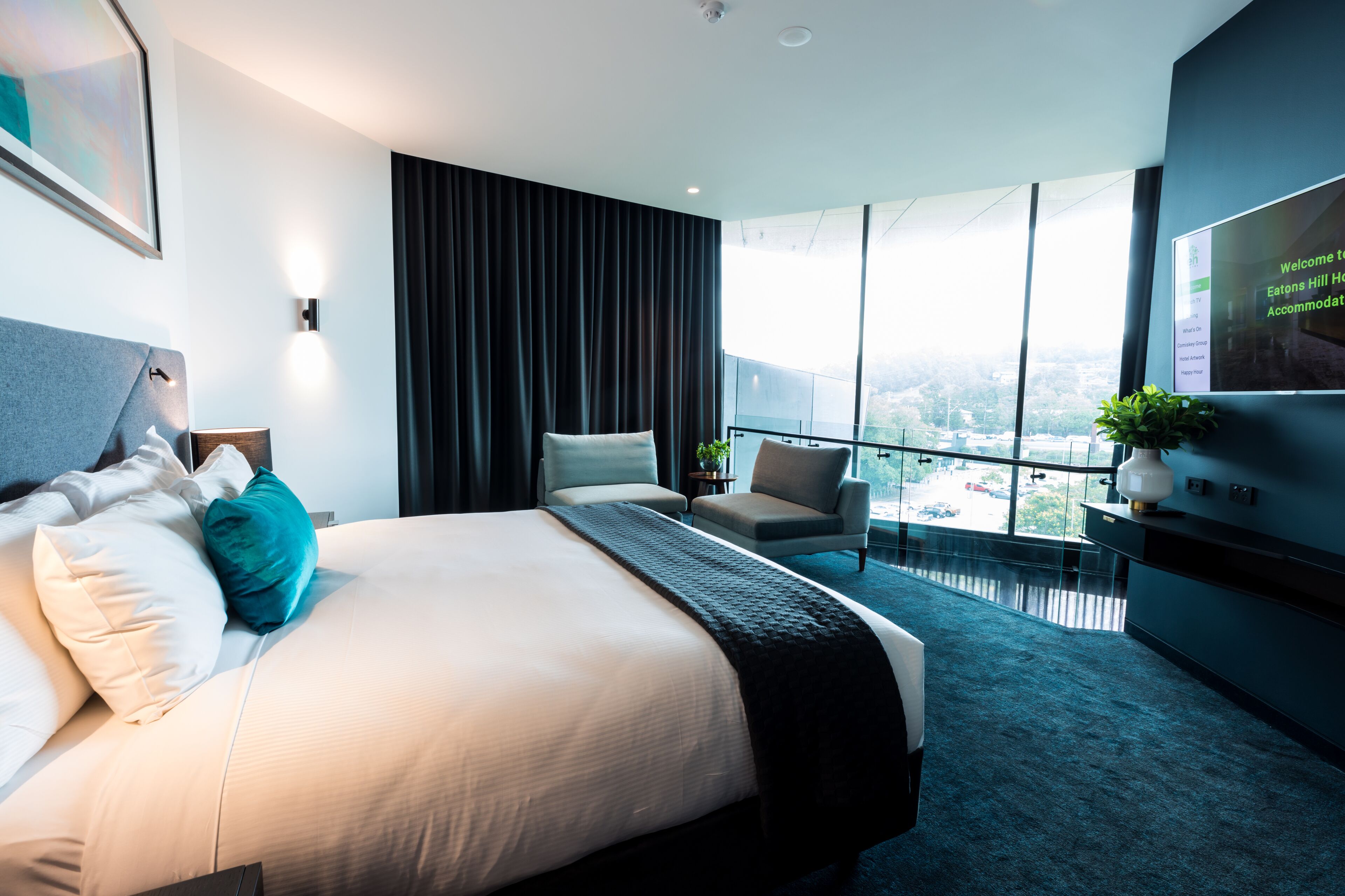 Una habitación de hotel moderna con una cama grande, una zona de estar con una silla y una mesita, un televisor y un balcón con vista a la ciudad.