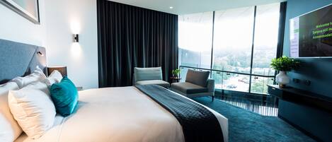 A modern hotel room with a large bed, a seating area with a chair and a small table, a TV, and a balcony with a city view.