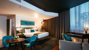 Una habitación de hotel moderna con una cama, dos sillones azules, un escritorio y un ventanal con cortinas.