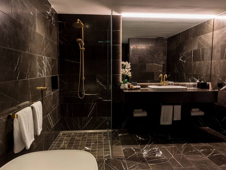 Ein modernes Badezimmer mit dunklen Marmorwänden, einer Glastrennwand zur Dusche, einer weißen Badewanne und einem Waschtisch mit Spüle und Handtuchhalter.
