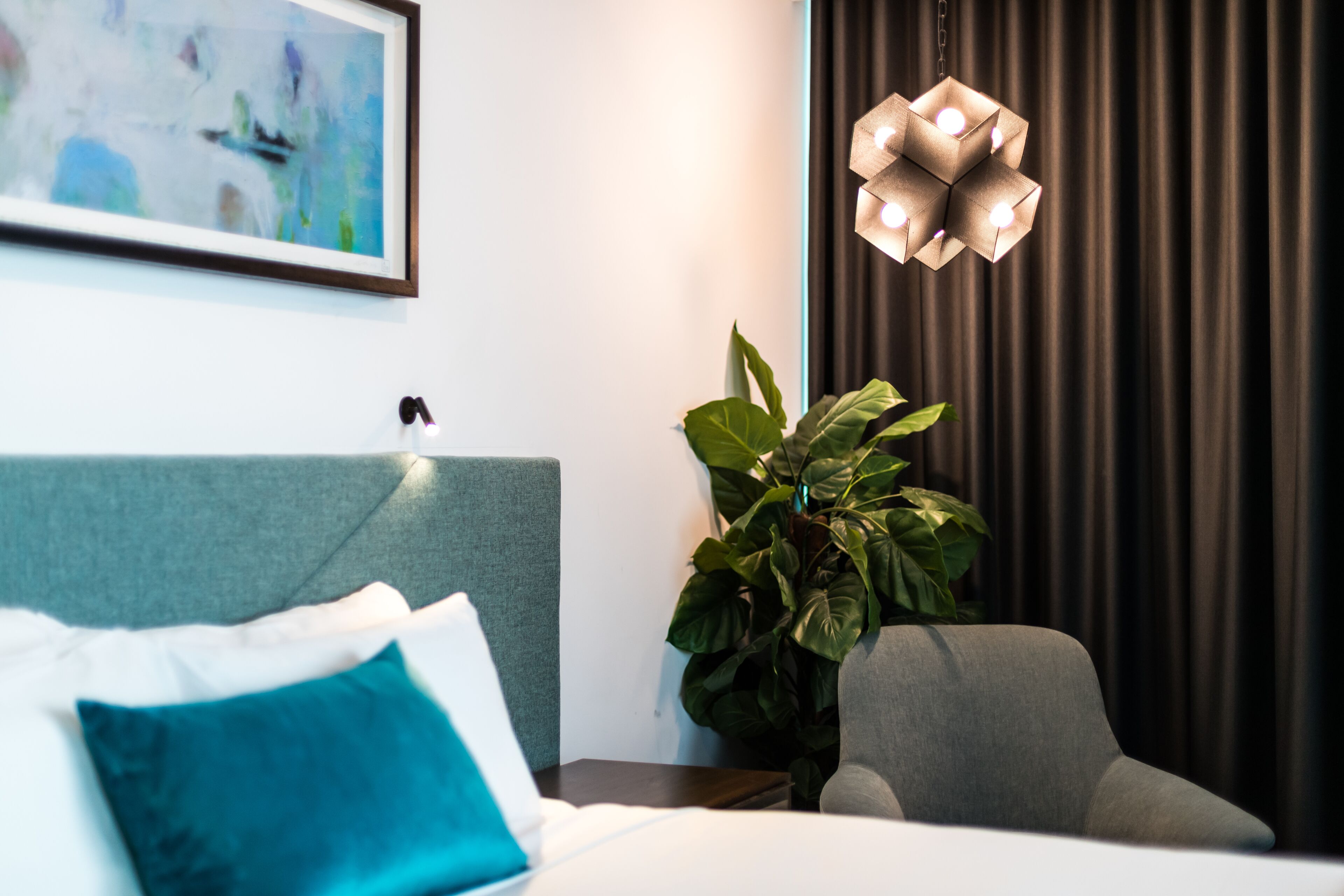 A hotel room with a bed, a grey armchair, a potted plant, a modern pendant light, and a framed abstract painting on the wall.