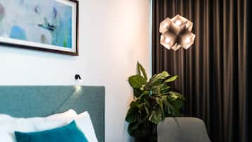A hotel room with a bed, a grey armchair, a potted plant, a modern pendant light, and a framed abstract painting on the wall.
