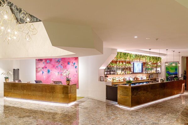 Un área de recepción moderna con piso de mármol, una mesa de recepción, una zona de bar con pared verde y un cuadro abstracto de gran tamaño.