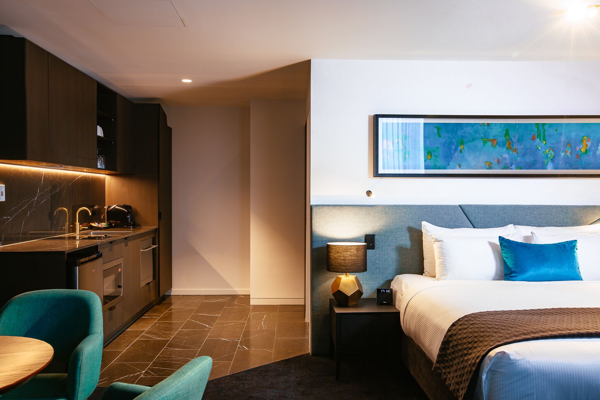 A hotel room with a bed, bedside table, lamp, and a painting on the wall.