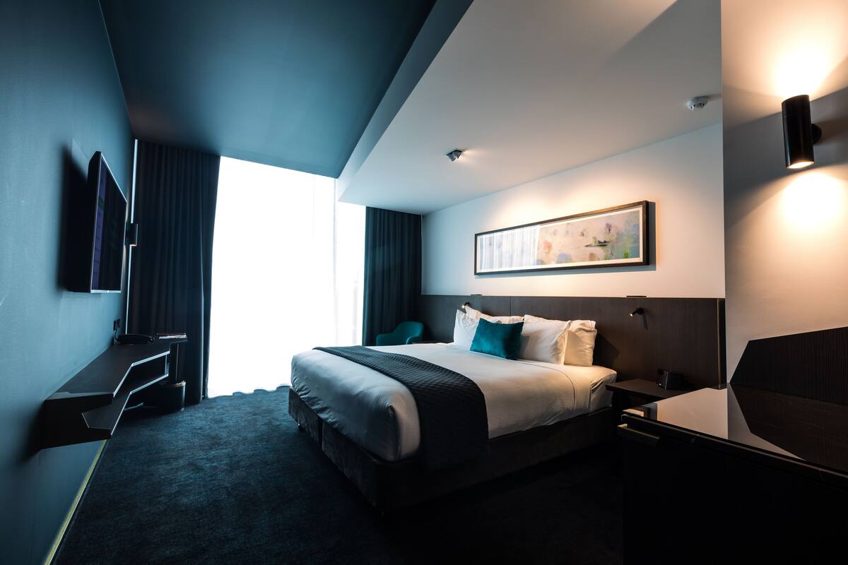 A modern hotel room with a large bed, a desk, a chair, and a flat-screen TV mounted on the wall.
