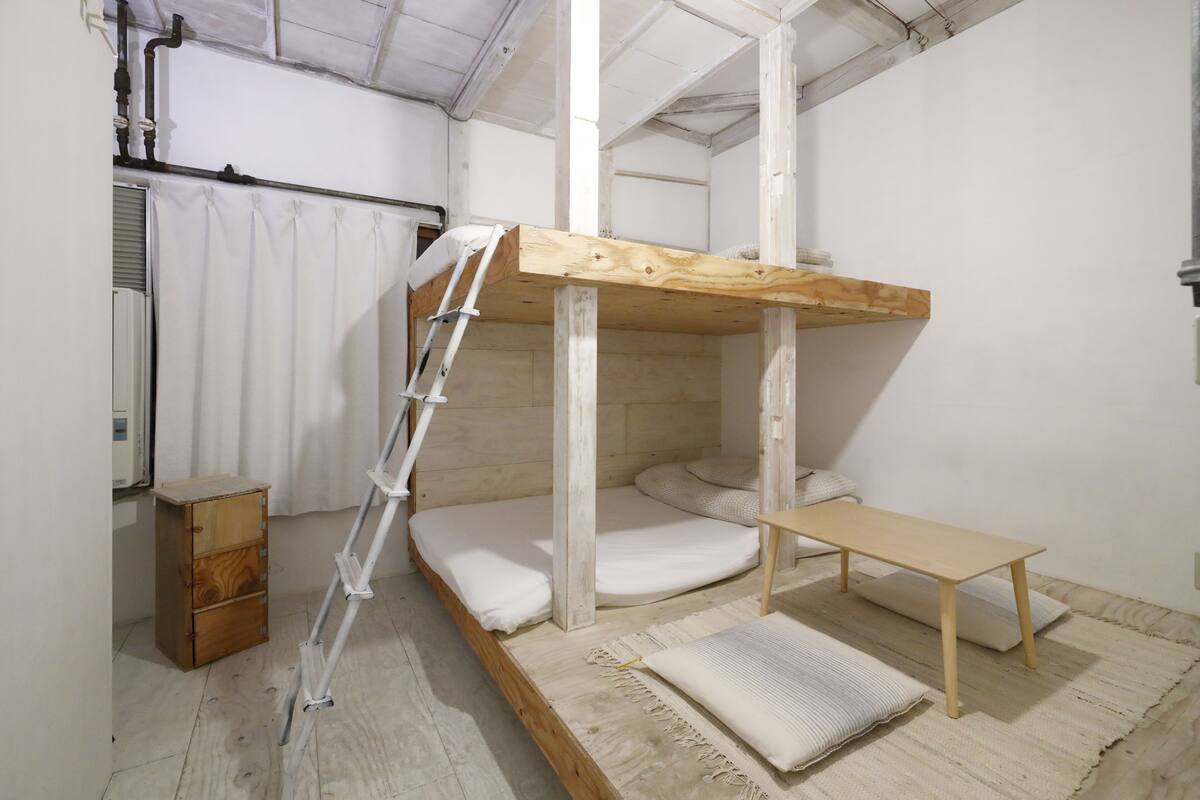 Ein Stockbett mit einer Leiter, ein kleiner Holzschrank und eine Bank in einem Raum mit weißen Wänden und Holzboden.
