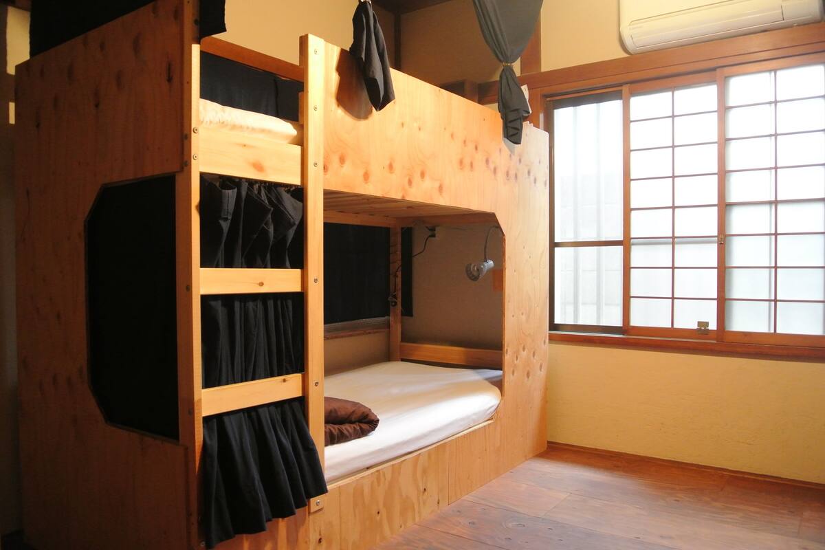 Ein Schlafzimmer mit Etagenbetten, Holzboden, einem Fenster mit Gitterfenstern und einem Vorhang auf der linken Seite.