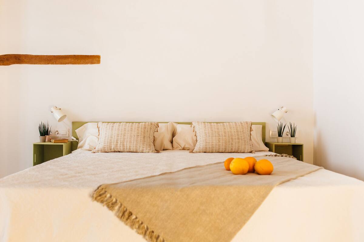 A neatly made bed with a beige blanket, two bedside tables with lamps, and a few oranges on the bed.