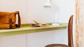 A room with a green shelf holding a brown bag and an open book, a white desk lamp, and a wicker chair.