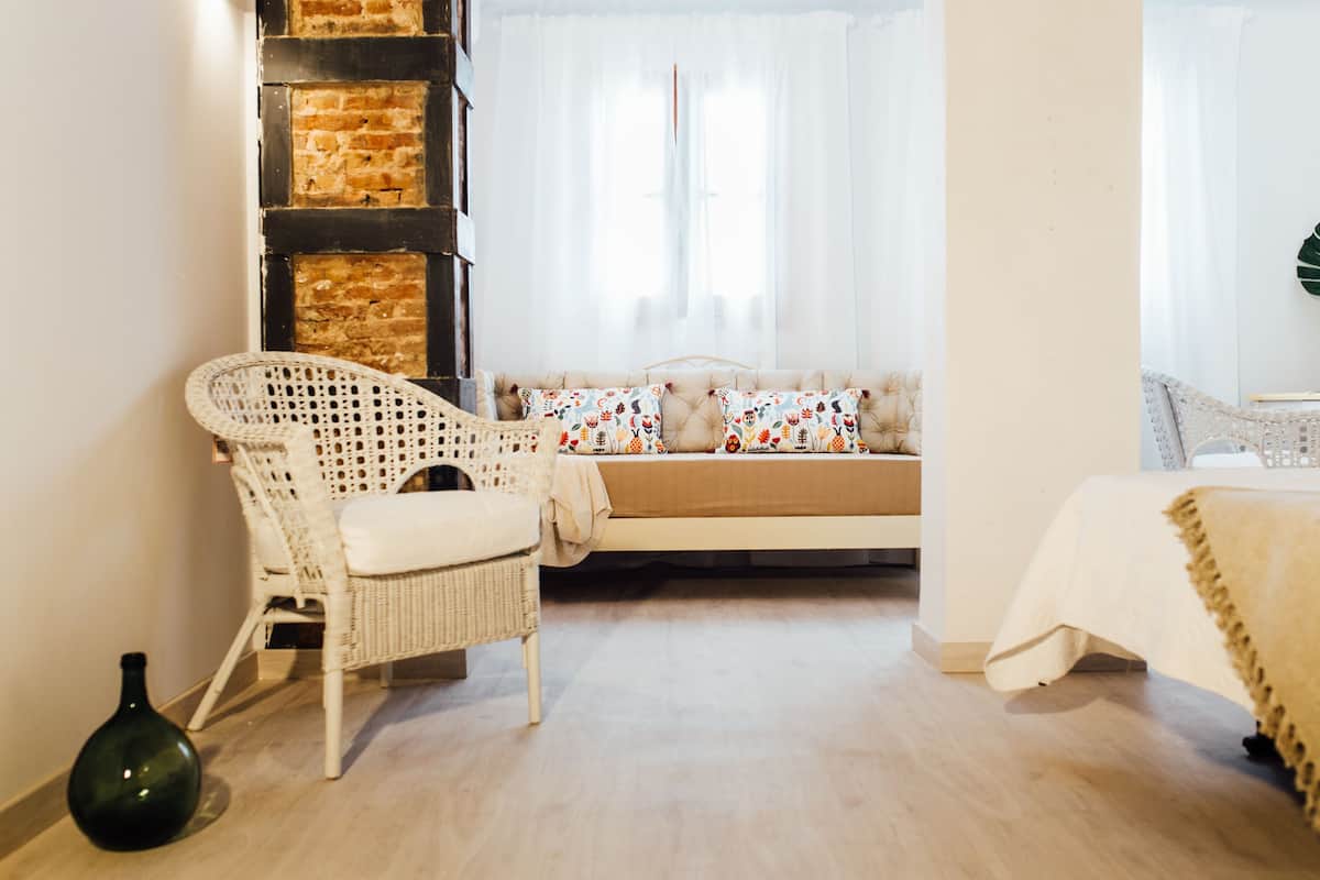 A room with a brick wall, a white wicker chair, a sofa with patterned cushions, and a bed with a beige cover.