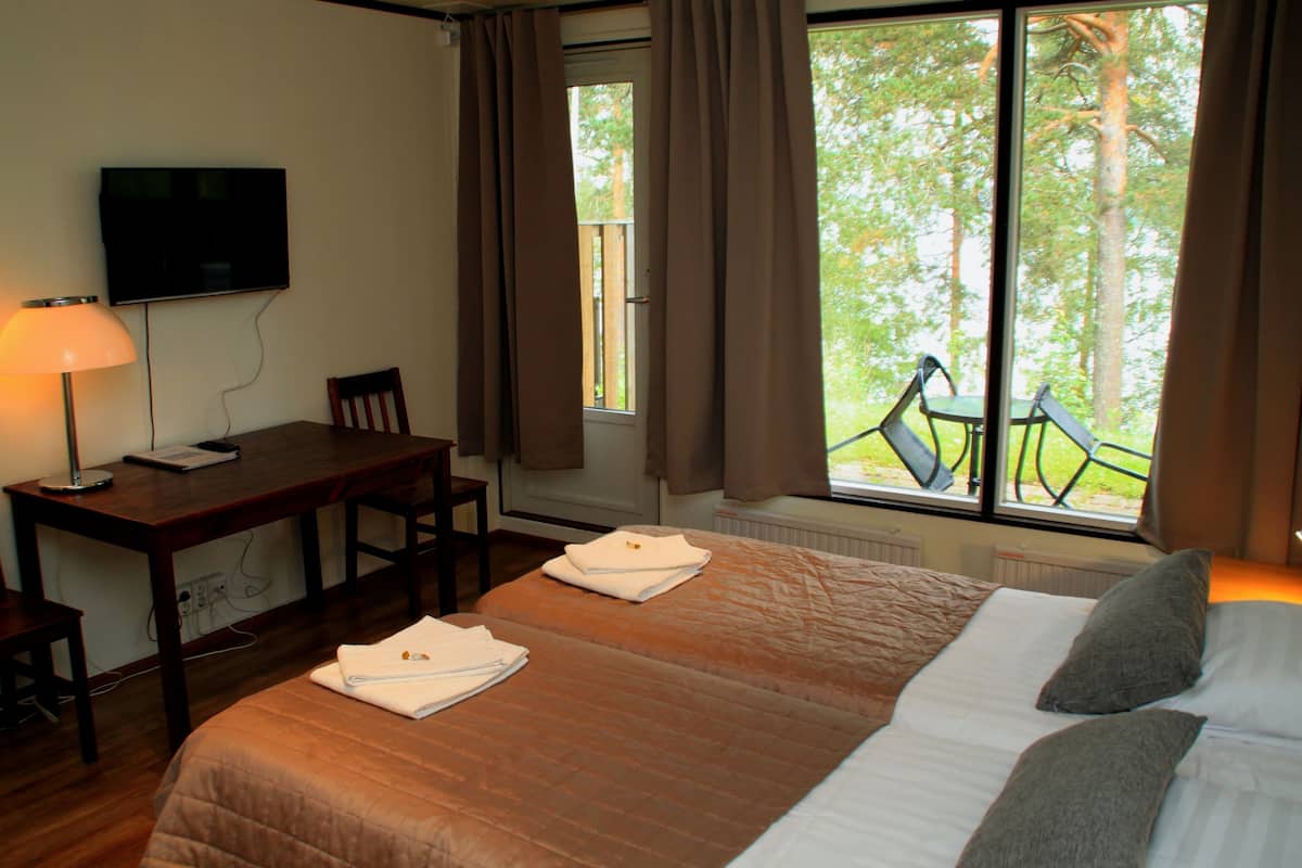 A hotel room with a bed, a desk, a chair, a TV, and a window with a view of trees and a balcony.