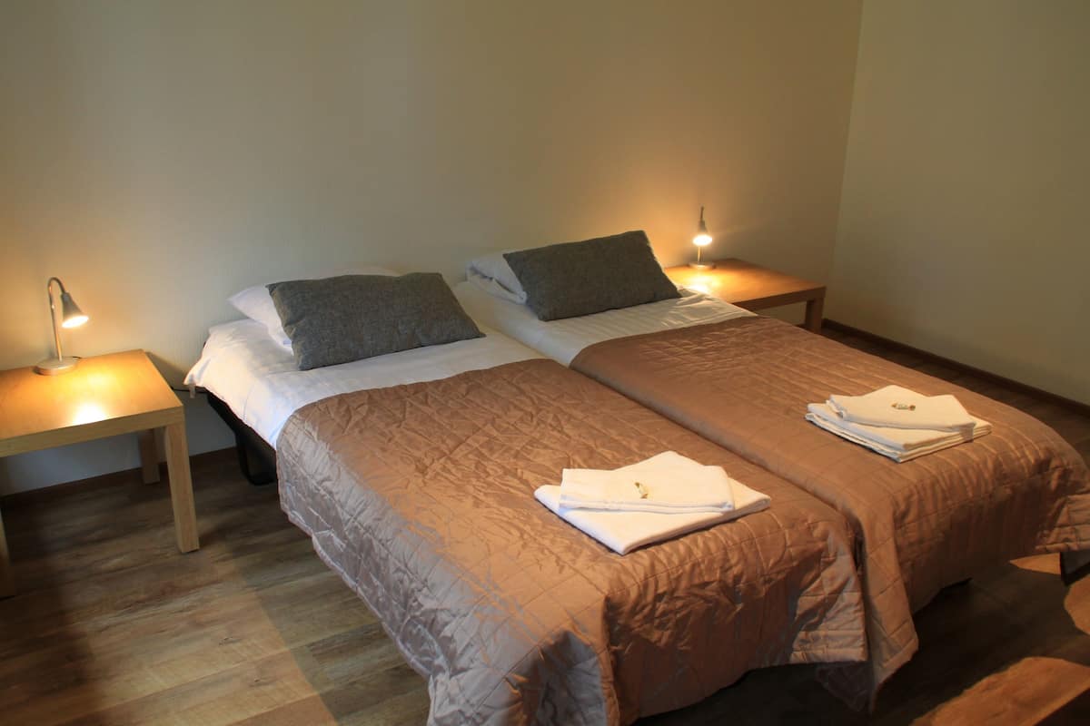 A double bed with a quilted brown cover, two pillows, and a folded towel on the bed. There are two bedside tables with lamps.