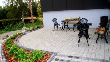 Une terrasse extérieure avec un sol en briques, entourée de verdure et d’un mur blanc.