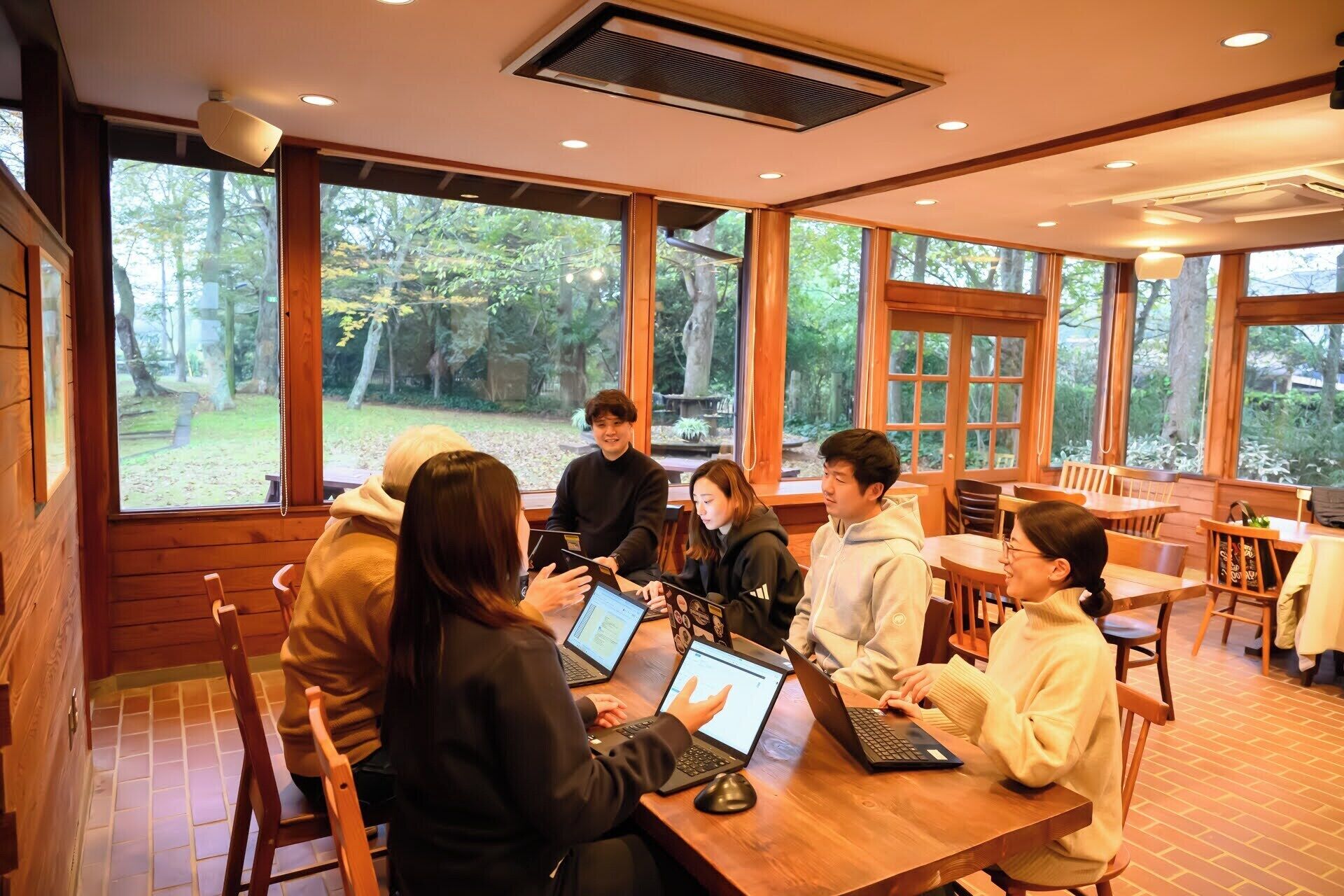 Un grupo de personas sentadas alrededor de una mesa de madera en una habitación con ventanas amplias, trabajando en portátiles.