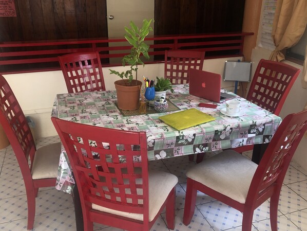 A dining area with a table covered by a floral tablecloth, surrounded by red chairs with cushioned seats. A potted plant is on the table, and a laptop is placed on the table's right side. - Yogi-Homestay (Koror)