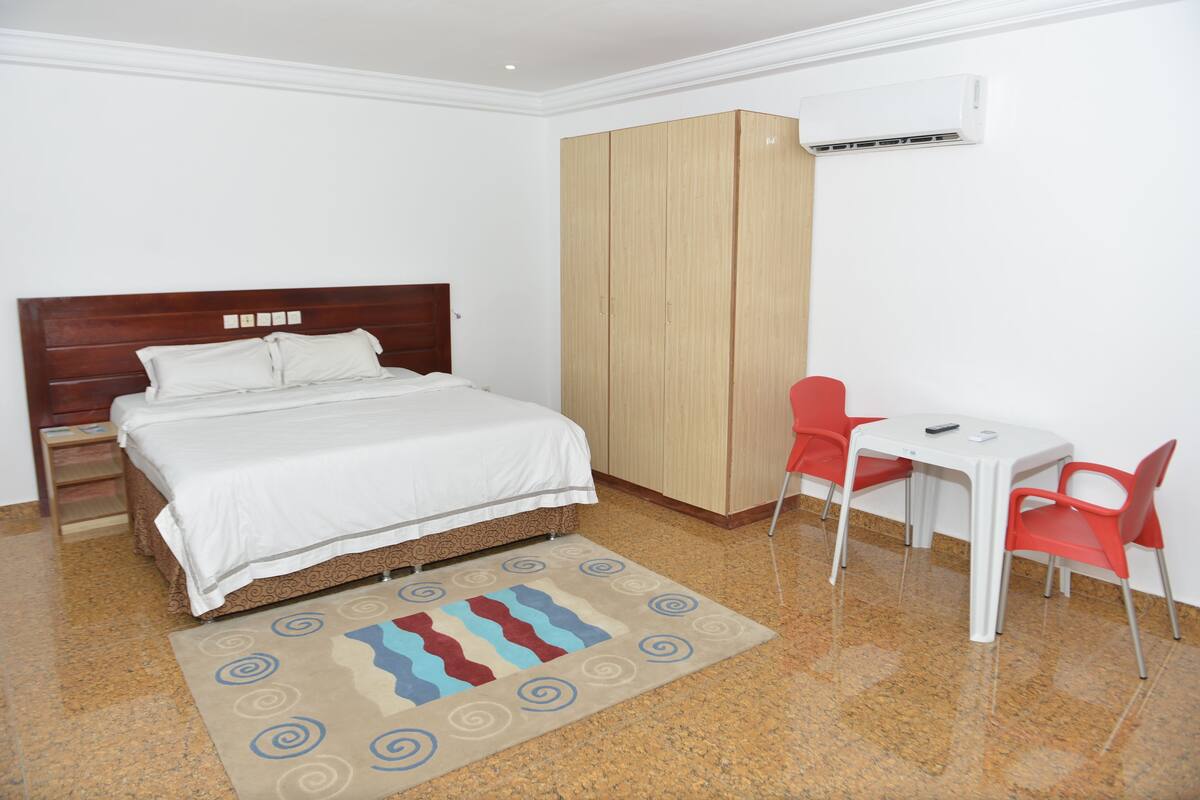 Una habitación de hotel con cama, armario, escritorio con sillas y aire acondicionado.