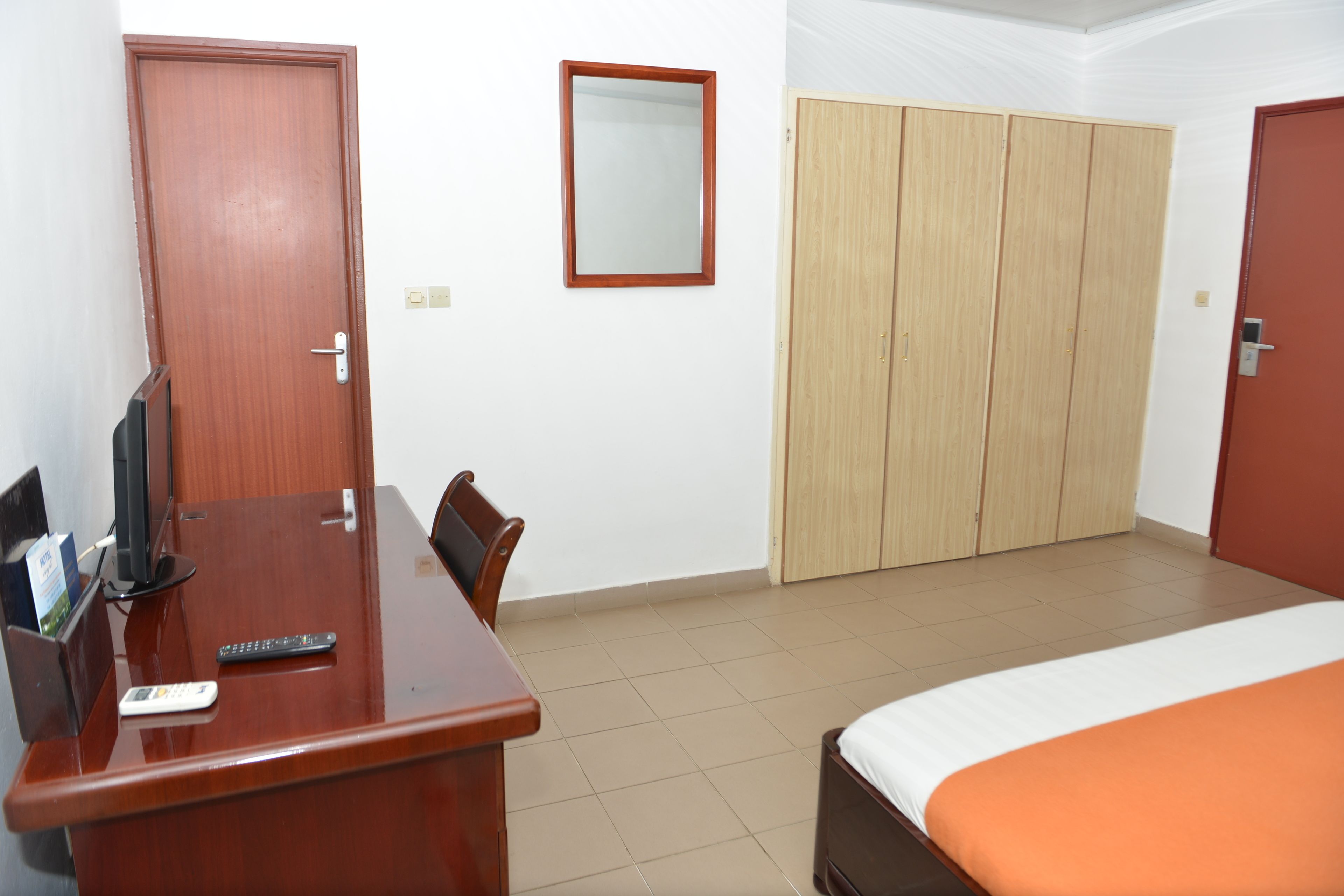 A hotel room with a wooden desk, a bed with an orange bedspread, a wardrobe, and a mirror on the wall.